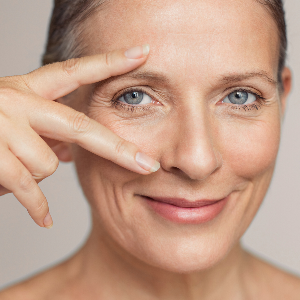 Mulher madura sorrindo suavemente, com pele saudável e toque leve próximo aos olhos, simbolizando cuidados com a região dos olhos.