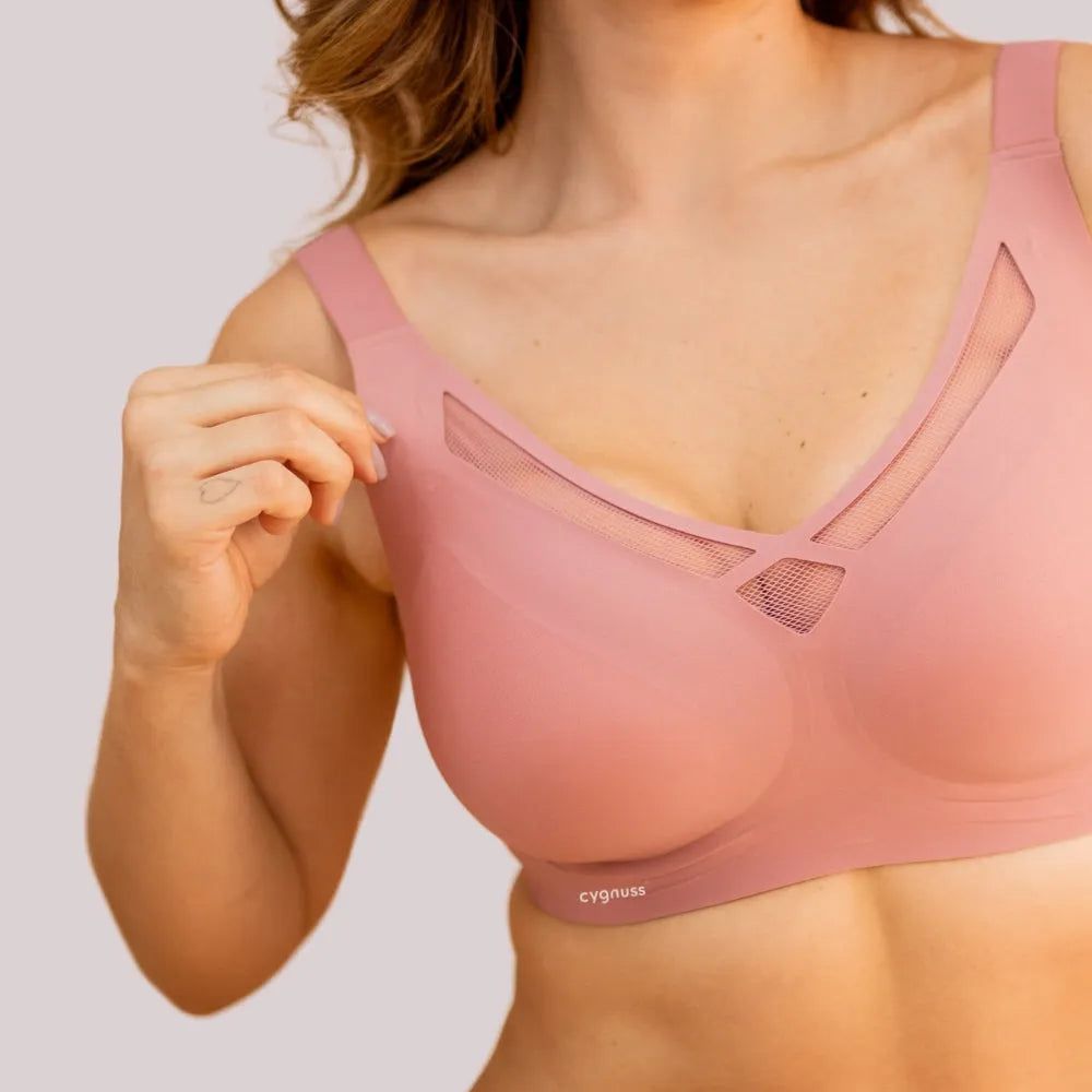 Close-up de uma mulher vestindo o Sutiã Dreams na cor rosa, destacando os detalhes vazados em tule no decote e as alças largas para maior sustentação. O logo da Cygnuss está visível na parte inferior do sutiã. A modelo segura delicadamente uma das alças, evidenciando o ajuste confortável do tecido sem costuras.