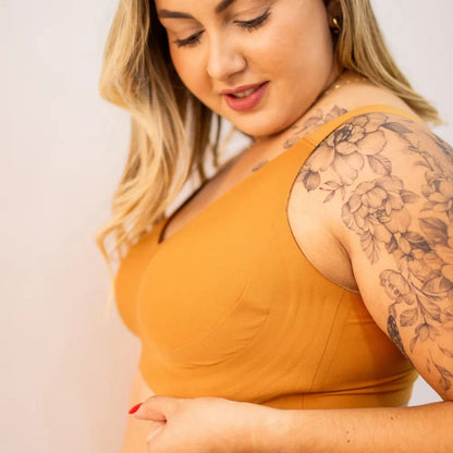 Mulher loira com tatuagem floral no ombro veste o Sutiã Curves Cygnuss na cor laranja, olhando para baixo enquanto sente o tecido macio e confortável.