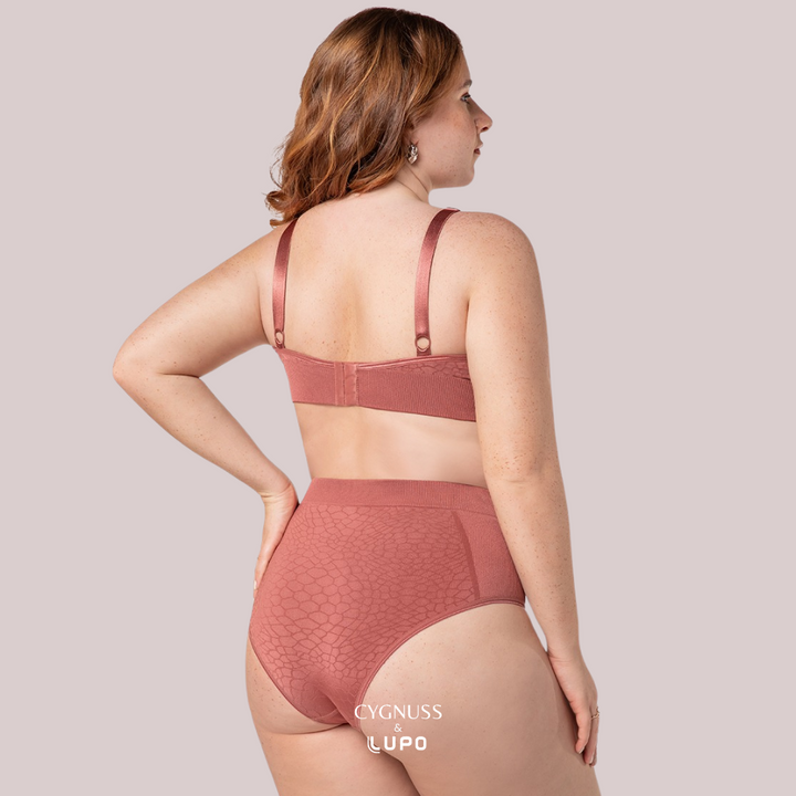 Vista traseira de uma mulher com cabelos ruivos usando um conjunto de lingerie na cor telha, composto por um sutiã e uma calcinha modeladora de cintura alta. O tecido texturizado sem costuras oferece suporte modelador e ajuste confortável. O fecho traseiro do sutiã garante melhor sustentação, enquanto a modelagem da calcinha realça a silhueta. O logotipo da Cygnuss & Lupo está sobreposto na parte inferior da peça. 