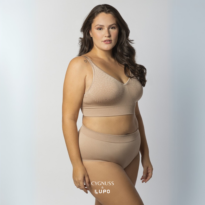 Mulher com corpo curvilíneo e cabelos longos usando um conjunto de lingerie nude sem costuras, composto por um sutiã modelador e uma calcinha de cintura alta. O tecido texturizado e elástico proporciona ajuste perfeito, conforto e sustentação. O logotipo da Cygnuss & Lupo está sobreposto na parte inferior da peça. Fundo branco com leve degradê azul.