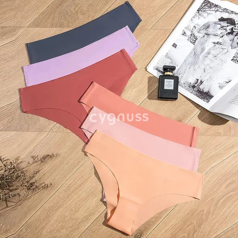 Imagem de calcinhas sem costura da Cygnuss dispostas sobre um fundo de madeira, apresentando tons suaves e quentes, como lilás, cinza, terracota, rosa e pêssego. Ao lado, um frasco de perfume e uma revista complementam a composição sofisticada.