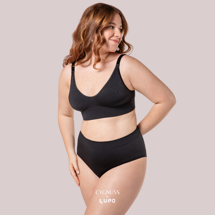 Mulher ruiva de cabelos cacheados usando um conjunto de lingerie preto sem costuras, composto por um sutiã modelador e uma calcinha de cintura alta. O tecido texturizado proporciona conforto, sustentação e um ajuste esculpido ao corpo. A modelo sorri levemente, transmitindo bem-estar e confiança. O logotipo da Cygnuss & Lupo está sobreposto na parte inferior da peça. Fundo nude rosado.
