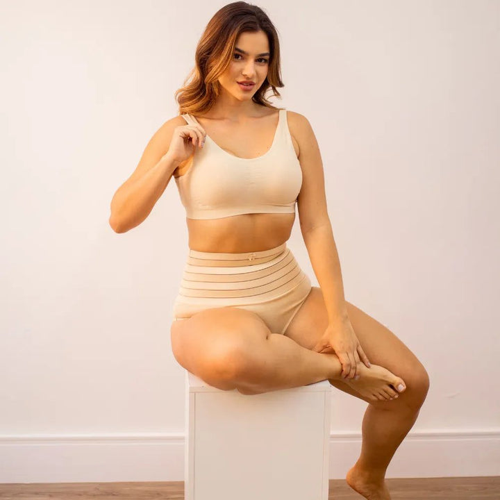 Imagem de uma mulher sentada, usando um conjunto de sutiã e calcinha modeladora Model - Seca Barriga, ambos na cor bege. A calcinha possui listras horizontais e uma flor delicada na faixa superior. Ela sorri e segura a alça do sutiã enquanto está em uma pose descontraída sobre um cubo branco.