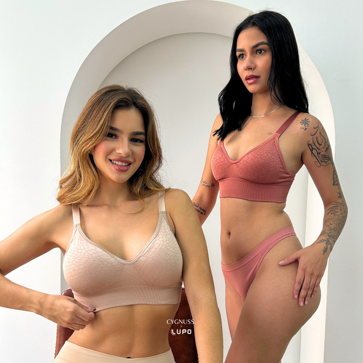 Duas mulheres usando os conjuntos Trend Curves Cygnuss x Lupo, uma na cor bege e a outra na cor terracota, em ambiente claro com arco decorativo, exibindo o sutiã com textura em relevo e calcinhas combinando.
