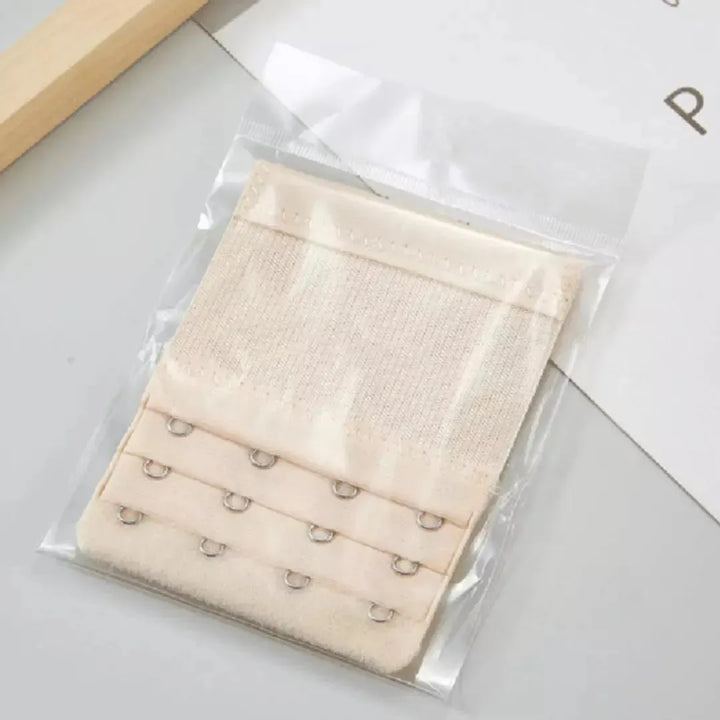 Extensor de sutia nude claro com tres niveis de ajuste e ganchos reforcados, embalado individualmente em plastico transparente. Acessorio ideal para aumentar o conforto e a adaptabilidade do sutia no dia a dia. Fundo claro com composicao minimalista.