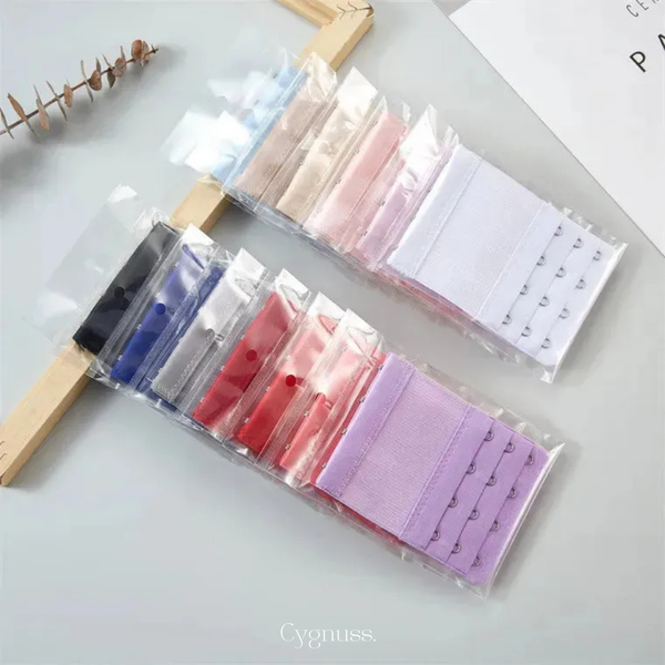 Extensores de sutia em diversas cores embalados individualmente, organizados sobre superfície clara. As cores incluem preto, azul, vermelho, rosa, lilas, branco e tons de nude. Cada extensor possui tres niveis de ajuste e ganchos metalicos, ideais para adaptar o sutia ao corpo com mais conforto.