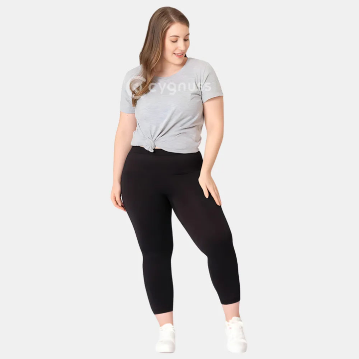 Mulher plus size usando a Legging Jade Fit Modeladora de Cós Alto na cor preta, combinada com camiseta cinza amarrada e tênis branco, em pose descontraída que evidencia o conforto e a versatilidade da peça no dia a dia.

