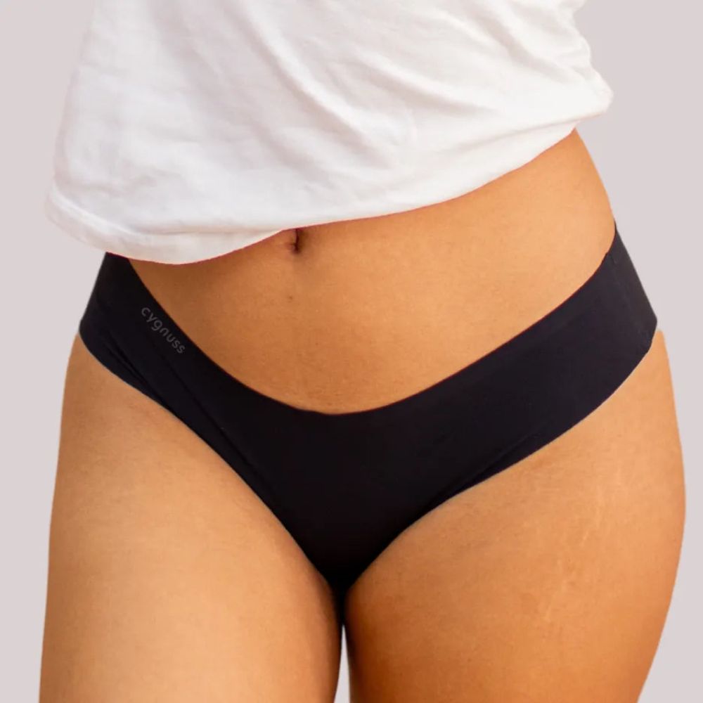 Close-up da região do quadril de uma mulher vestindo uma calcinha fio-dental preta sem costura da Cygnuss, combinada com uma camiseta branca. A imagem destaca o ajuste perfeito e o conforto do tecido.