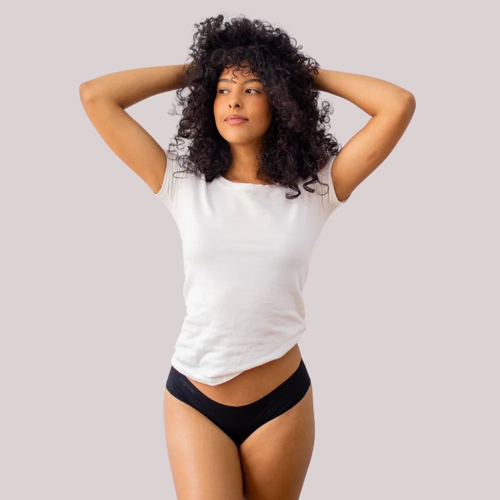 Mulher de cabelos cacheados posando com as mãos no cabelo, vestindo uma camiseta branca e uma calcinha fio-dental preta sem costura da Cygnuss. A imagem destaca o ajuste confortável e a elegância minimalista da peça