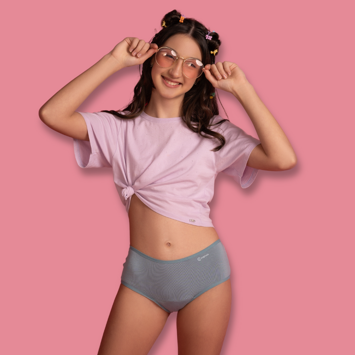 Modelo adolescente usando calcinha azul claro da linha ciclo teen com camiseta lilás e óculos estilosos, posando diante de fundo rosa, representando conforto e estilo jovem.