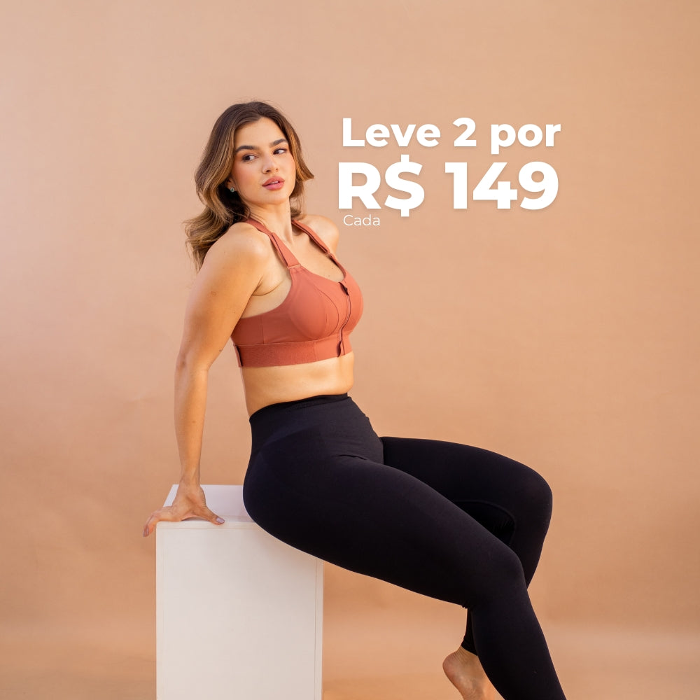 Mulher vestindo sutiã esportivo com zíper frontal Cygnuss em tom terroso e calça legging preta, sentada em um cubo branco, com fundo bege. Texto promocional indicando a oferta de "Leve 2 por R$ 149 cada