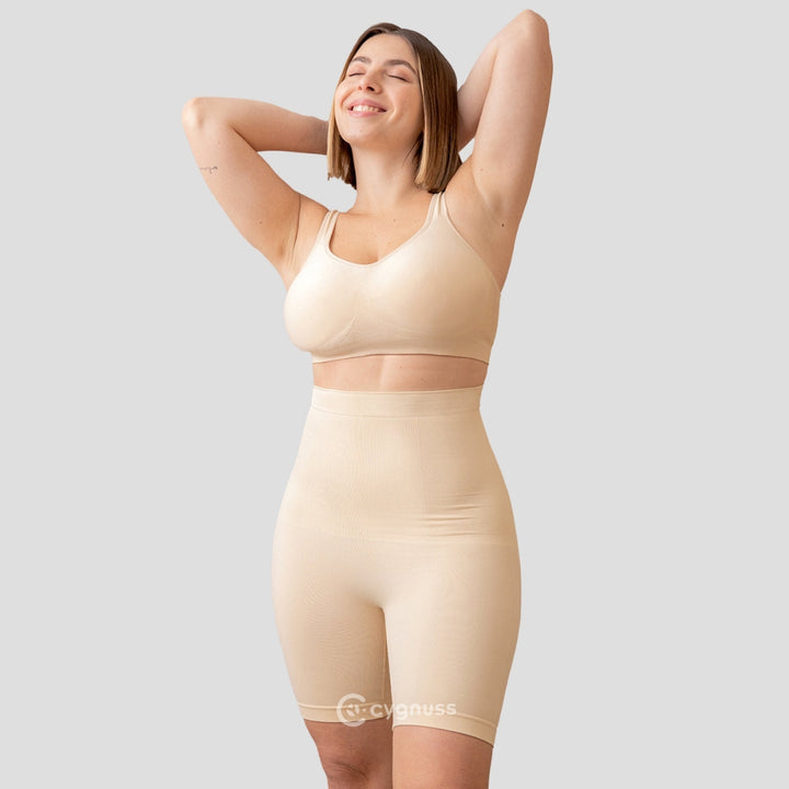 Mulher sorridente, de pé, usando o Short Premium Modelador com Faixas de Silicone na cor bege. O modelador proporciona um contorno perfeito ao corpo, com alta elasticidade e conforto, destacando a silhueta de forma elegante.
