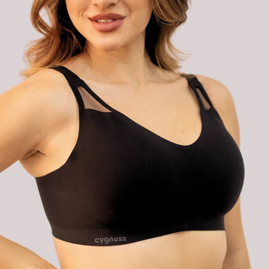 Mulher vestindo o Sutiã Academy na cor preta, destacando o design sem costuras, as alças largas e os detalhes vazados em tule no decote. A peça proporciona conforto e sustentação, com modelagem anatômica. O logo da Cygnuss está visível na parte inferior do sutiã.