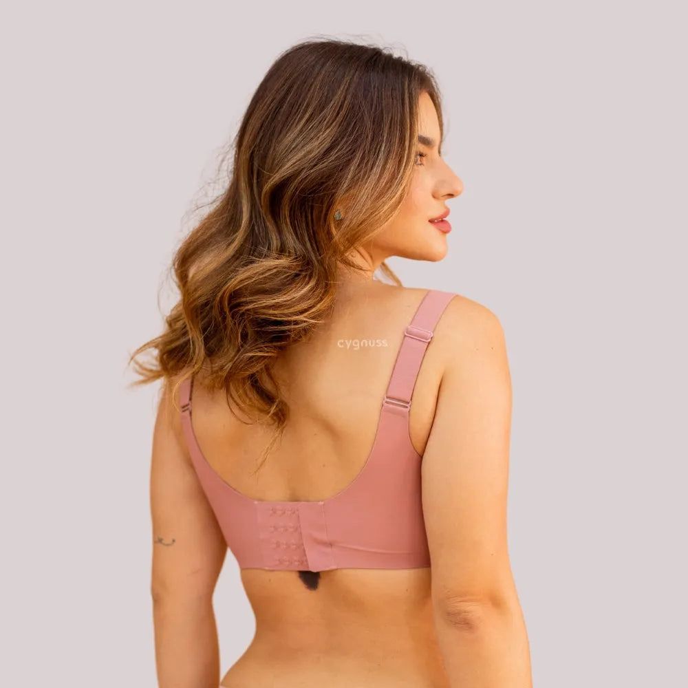 Mulher de costas vestindo o Sutiã Dreams na cor rosa, destacando o fecho ajustável com quatro níveis de regulagem e as alças largas para maior sustentação e conforto. O tecido sem costuras proporciona um ajuste suave ao corpo. O cabelo da modelo está solto e ondulado, e o fundo da imagem é em tom neutro.