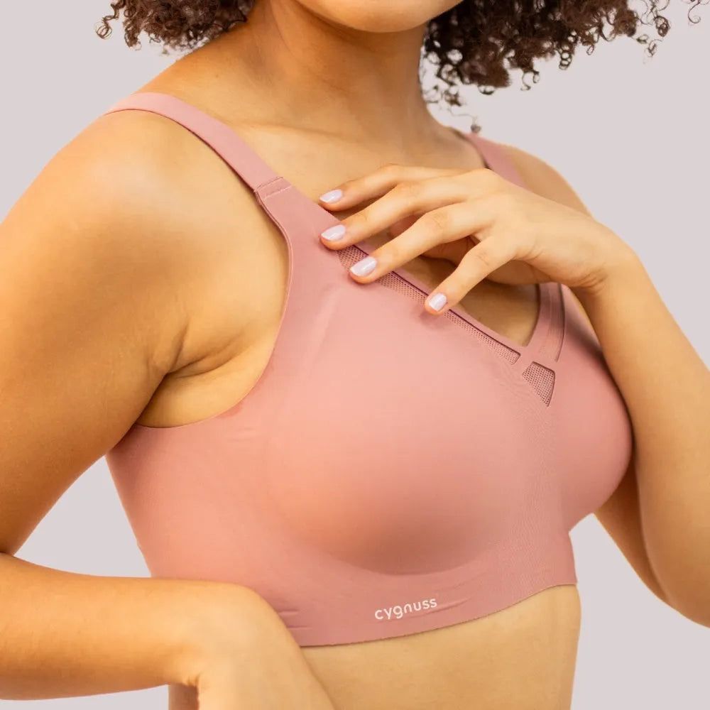 Close-up de uma mulher vestindo o Sutiã Dreams na cor rosé, destacando o design sem costuras, os detalhes vazados em tule no decote e as alças largas para maior conforto e sustentação. A modelo toca suavemente no sutiã, evidenciando a textura e ajuste da peça. O logo da Cygnuss está visível na parte inferior do sutiã.