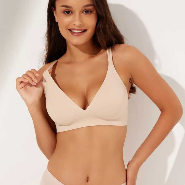 Mulher sorrindo, vestindo o Sutiã FIT bege com alças finas e bojo removível. Ela segura delicadamente uma das alças, destacando o decote em V e o caimento perfeito da peça com acabamento sem costura, ideal para o dia a dia com conforto e leveza.