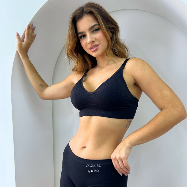 Mulher usando o Sutiã Trend Curves Cygnuss x Lupo na cor preta, com textura em relevo e decote em V, em pose frontal descontraída ao lado de uma parede branca com arco decorativo.

